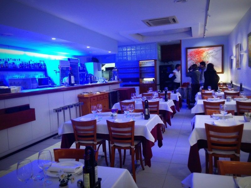 Un interior de restaurante poco iluminado con iluminación ambiental azul, mesas con manteles blancos, sillas de madera y un bar en la parte trasera.