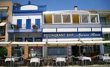 Un restaurante Bar María Rosa iluminado por el sol, con edificios blancos, molduras azules y mesas al aire libre colocadas bajo un toldo retráctil.
