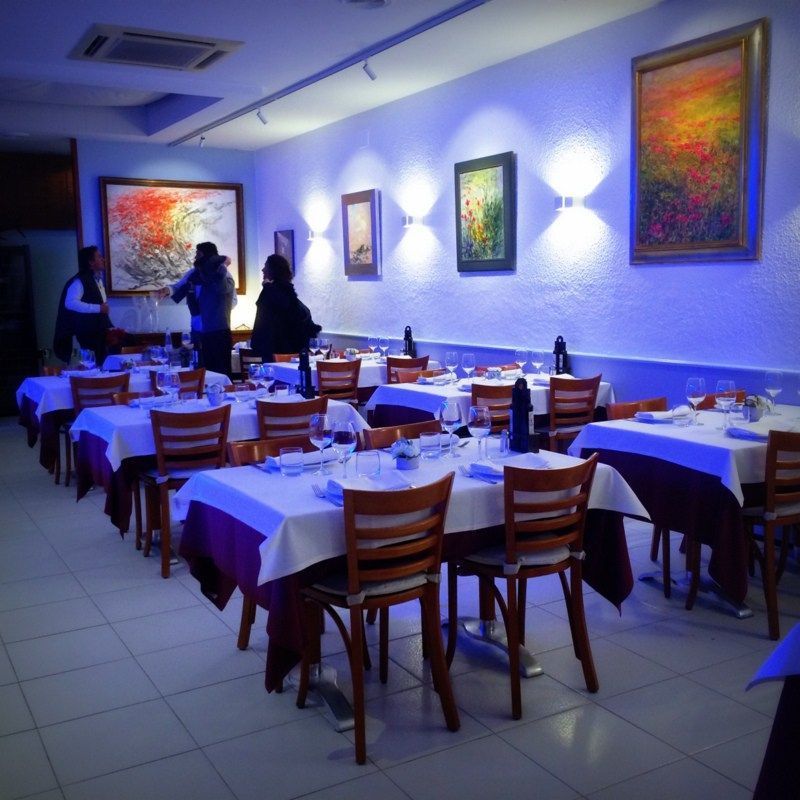 Un interior de restaurante con mesas cubiertas con manteles blancos, sillas de madera, iluminación ambiental azul y arte en una pared blanca.