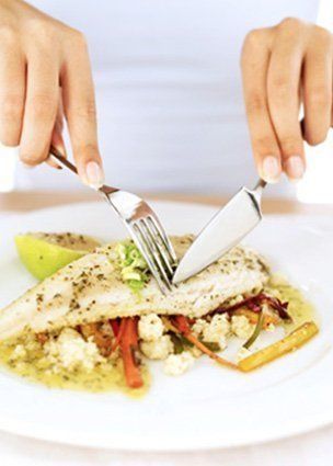 Las manos usan un tenedor y un cuchillo para cortar un filete de pescado sazonado servido sobre una cama de verduras y salsa en un plato blanco.