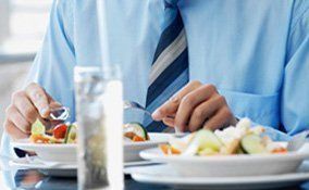 Una persona con camisa azul y corbata a rayas come una ensalada fresca en una mesa con un vaso de agua cerca.