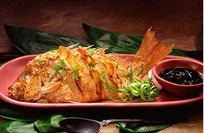 Un pescado frito entero adornado con cebollas verdes en un plato rojo, servido con un tazón pequeño de salsa oscura para mojar.