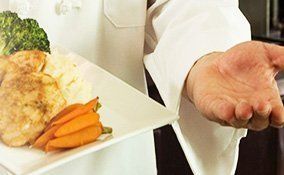 Un chef con uniforme blanco sostiene un plato de pollo, brócoli y zanahorias y extiende la otra mano hacia adelante.