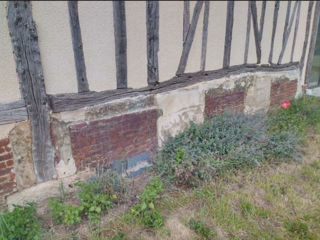 Vieux mur à colombages avec soubassement en briques et herbes hautes envahissantes au pied du bâtiment
