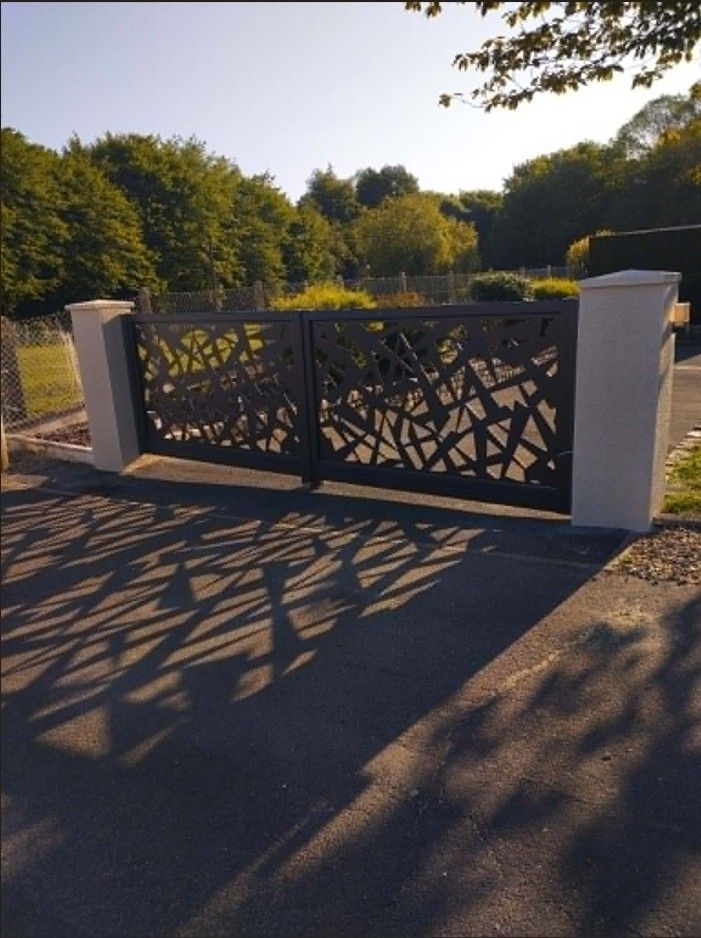Portail décoratif en métal noir avec des piliers blancs, au bord d'une route ensoleillée bordée d'arbres projetant de longues ombres.