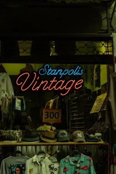 Letreiro de néon com a inscrição “Stamplis Vintage” acima da vitrine de uma loja de roupas com camisas e chapéus pendurados.
