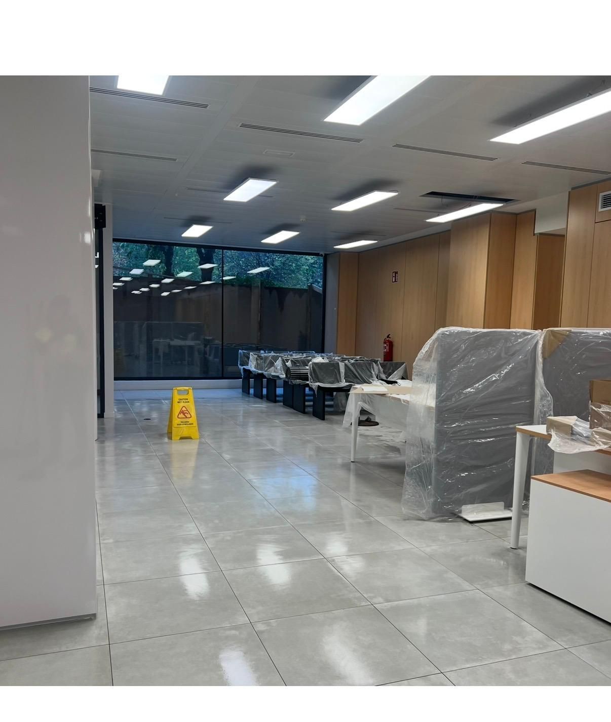 Un espacio de oficina vacío, recientemente renovado, con escritorios cubiertos de plástico y un cartel de piso mojado.