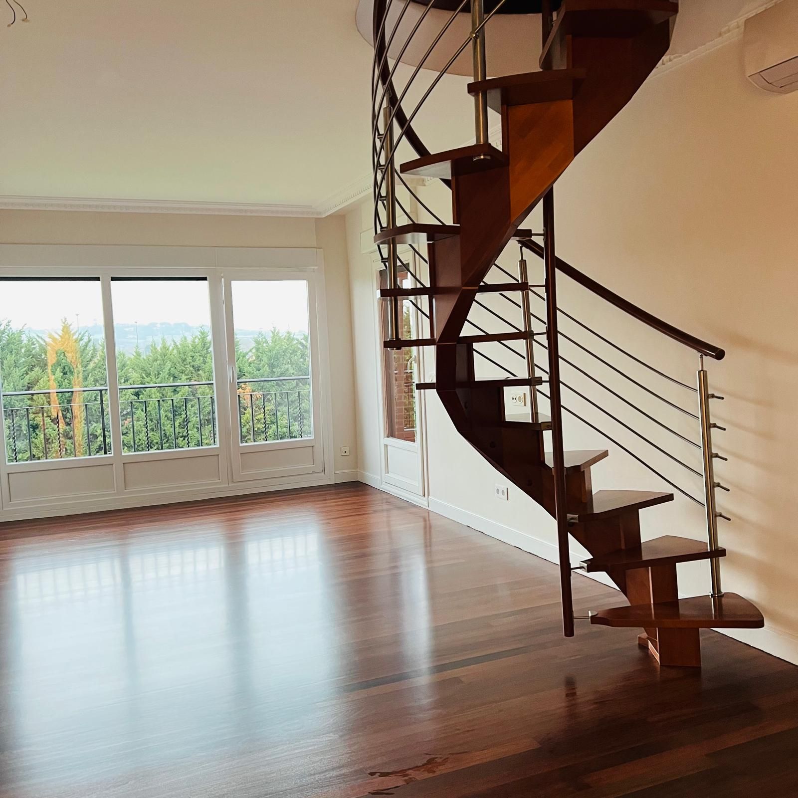 Escalera de caracol de madera en una habitación con grandes ventanales con vistas a los árboles y suelo de madera.