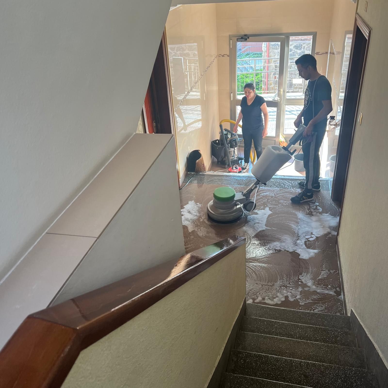 Personas limpiando una escalera de azulejos con una pulidora y una solución limpiadora; puerta de entrada visible.