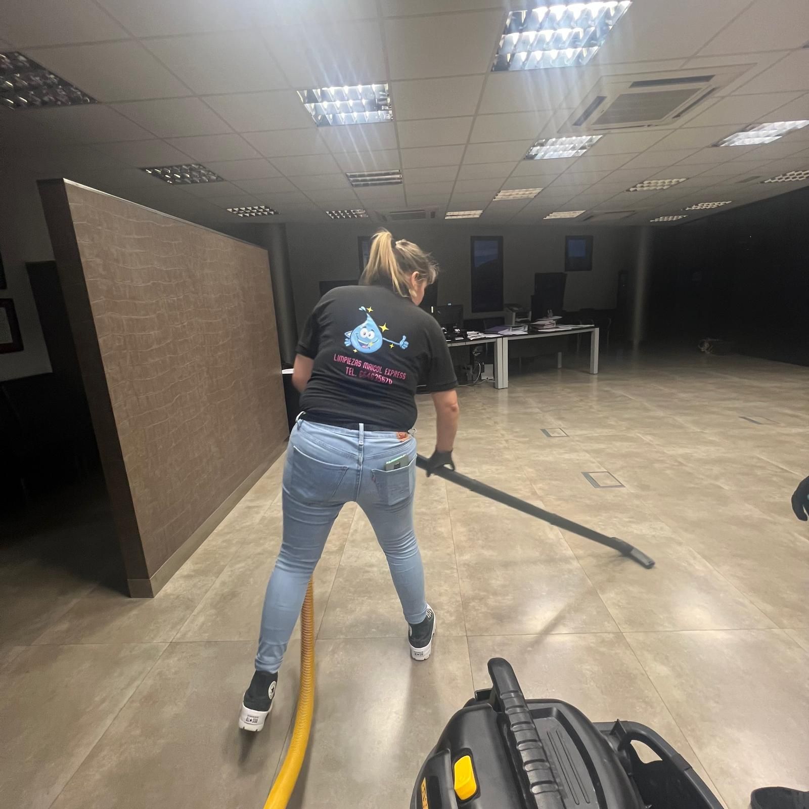 Mujer aspirando una gran pared marrón en una oficina, vestida con jeans y una camiseta negra, con la aspiradora visible.