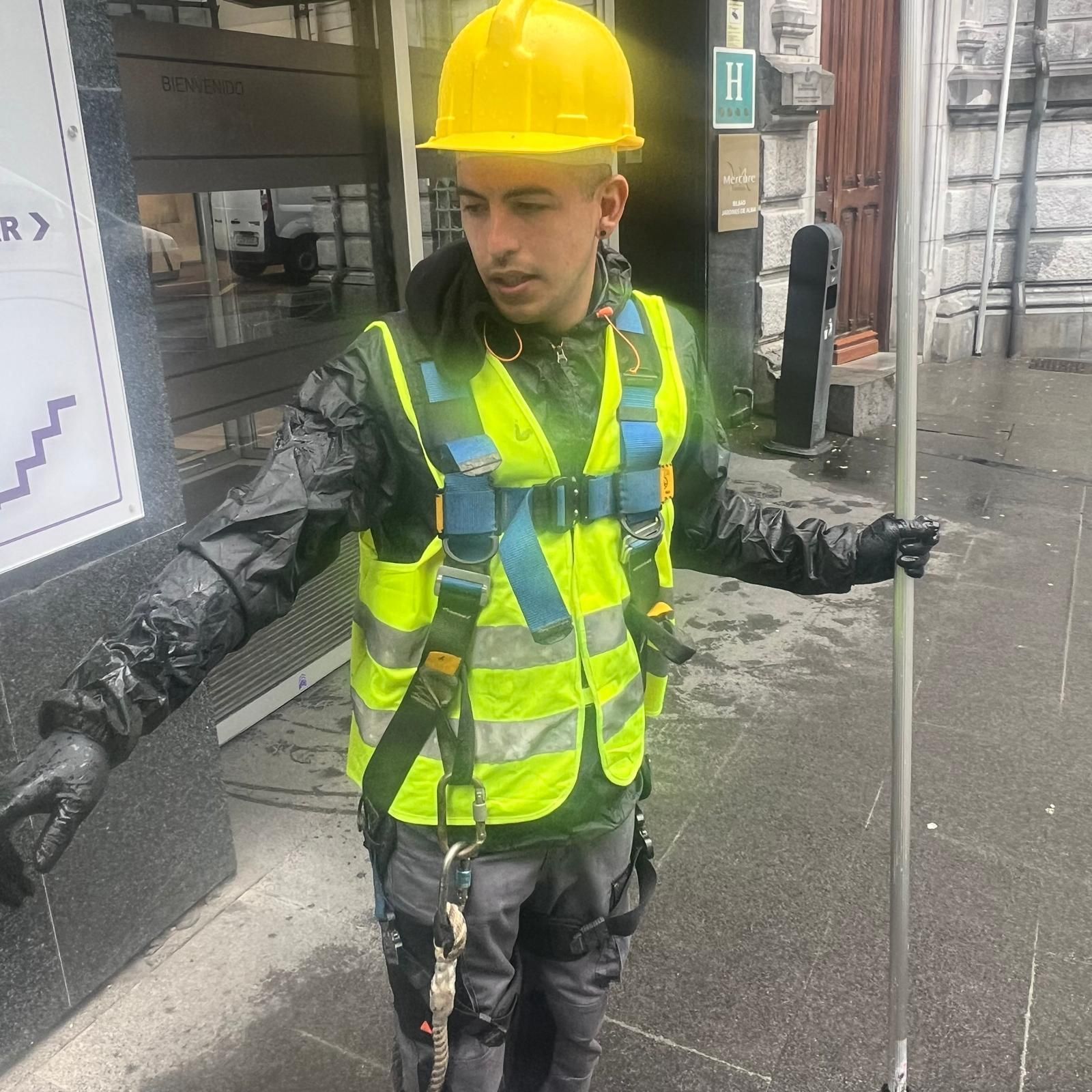Hombre con casco amarillo y chaleco de seguridad sosteniendo un poste, de pie afuera de un edificio.