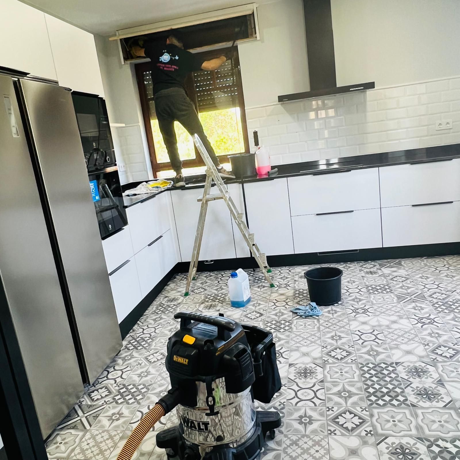 Persona limpiando una ventana de la cocina, de pie sobre una escalera. Cocina en blanco y negro con suelo estampado.