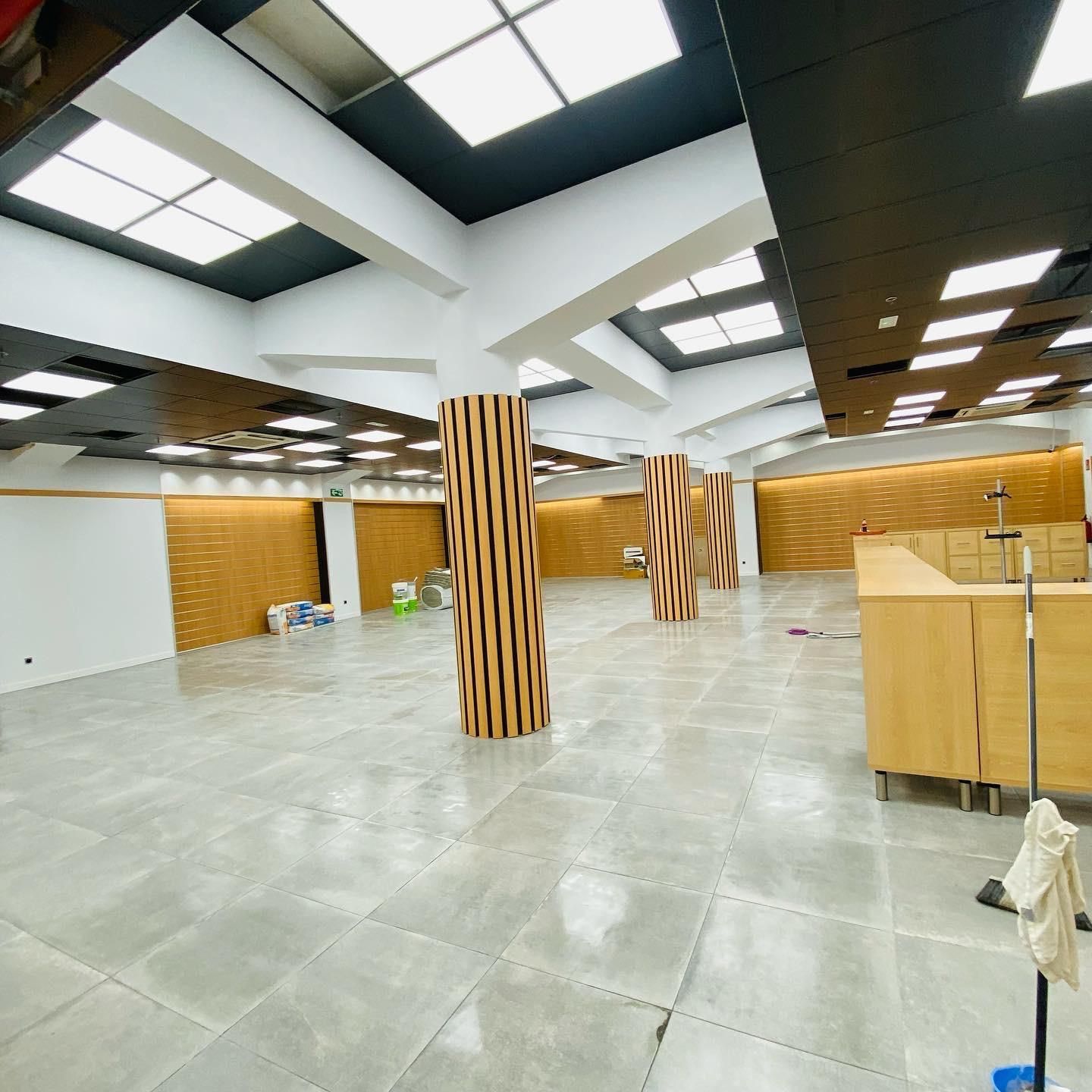 Espacio comercial vacío con paredes de colores claros, detalles de techo oscuros, columnas de madera y pisos de baldosas.