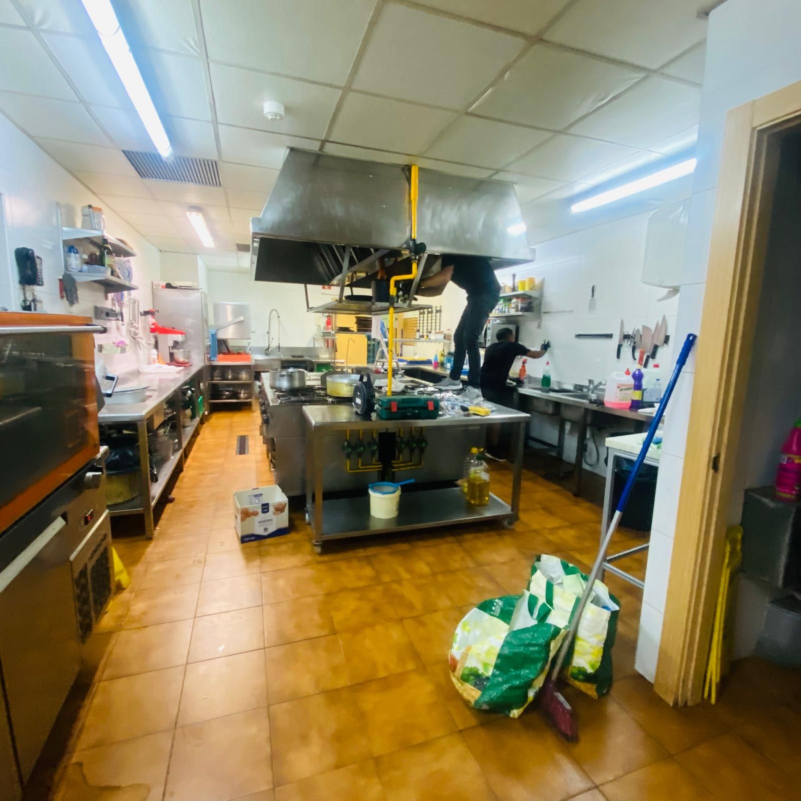 Dos personas trabajando en una cocina comercial, con una gran campana extractora sobre una isla de acero inoxidable.