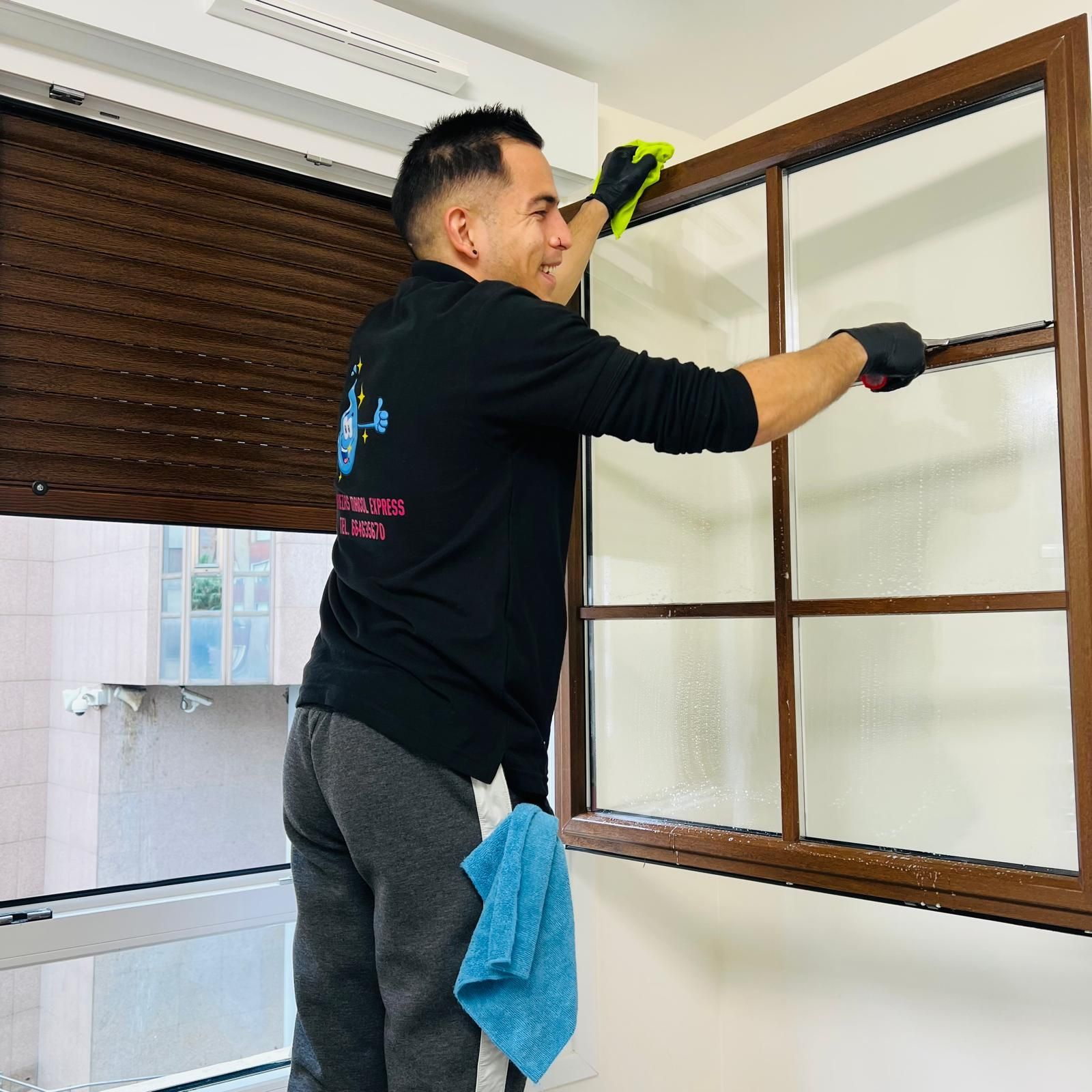 Hombre limpiando una ventana, con guantes negros, en una habitación con marcos de ventanas marrones y una persiana marrón.