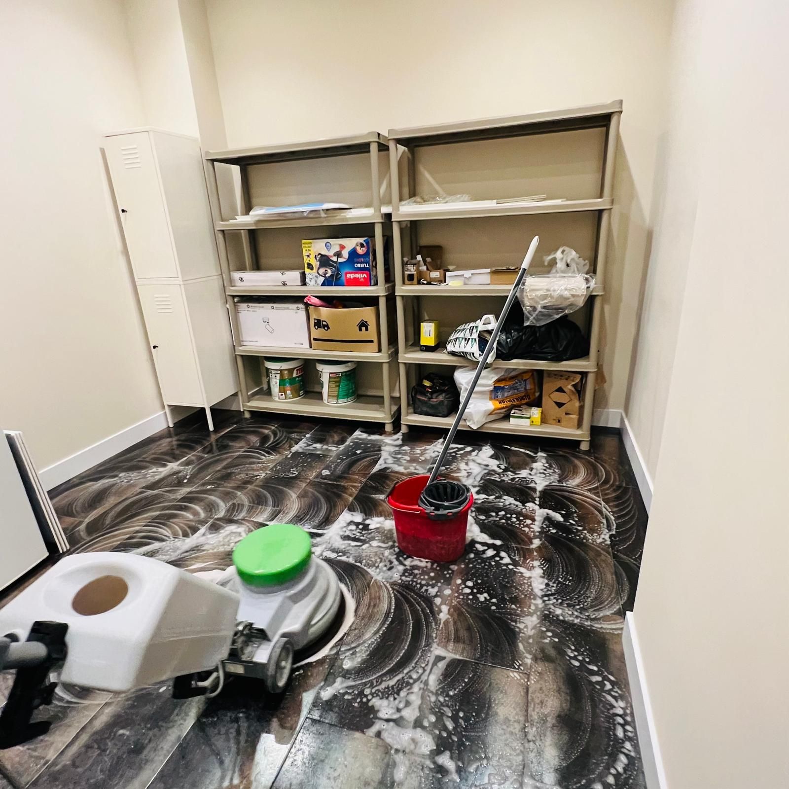 Limpieza de suelo en curso con máquina, estanterías y taquilla en habitación pequeña.