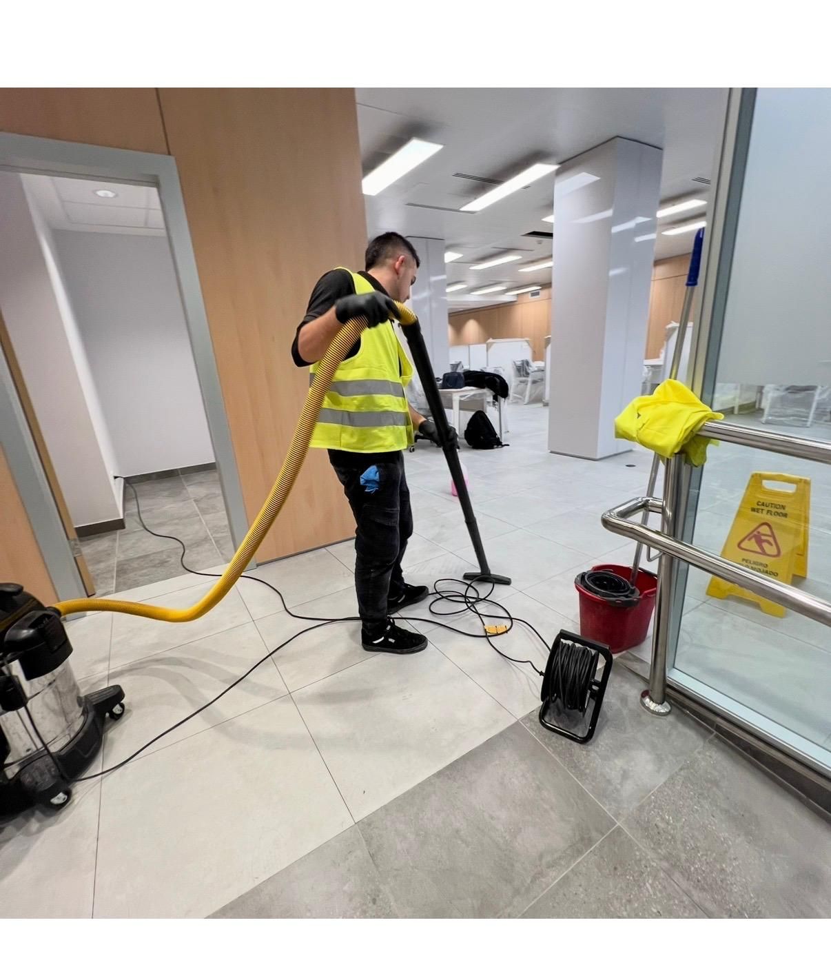 Hombre con chaleco amarillo aspirando un espacio comercial, limpiando, cerca de un cartel de piso mojado.