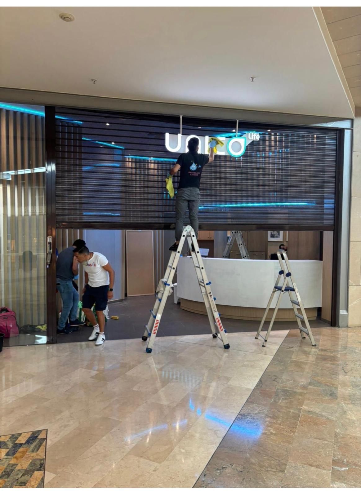Trabajadores instalando o limpiando un letrero en un centro comercial. Uno en una escalera, otros cerca.