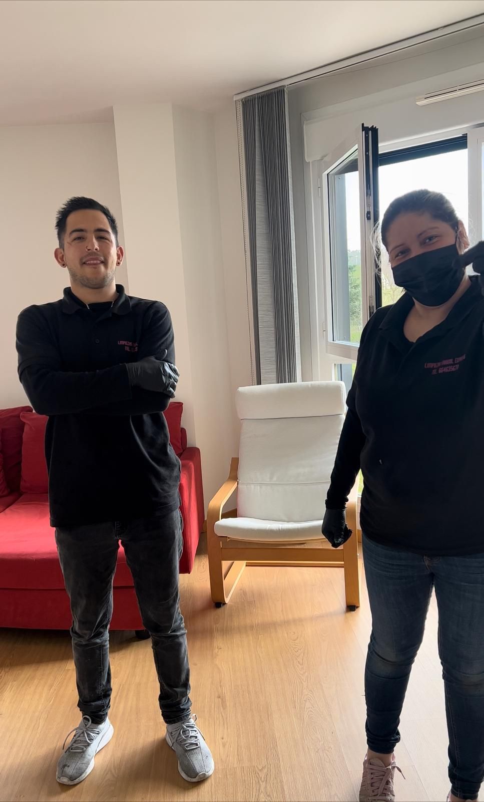 Dos personas, una con mascarilla, están en un interior. Un hombre con los brazos cruzados sonríe, mientras que otra persona levanta un brazo, con guantes.