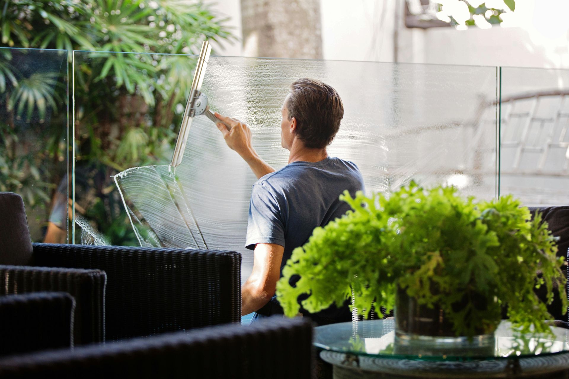Hombre limpiando una ventana con una escobilla de goma al aire libre. En la escena se ven plantas verdes y muebles de mimbre.