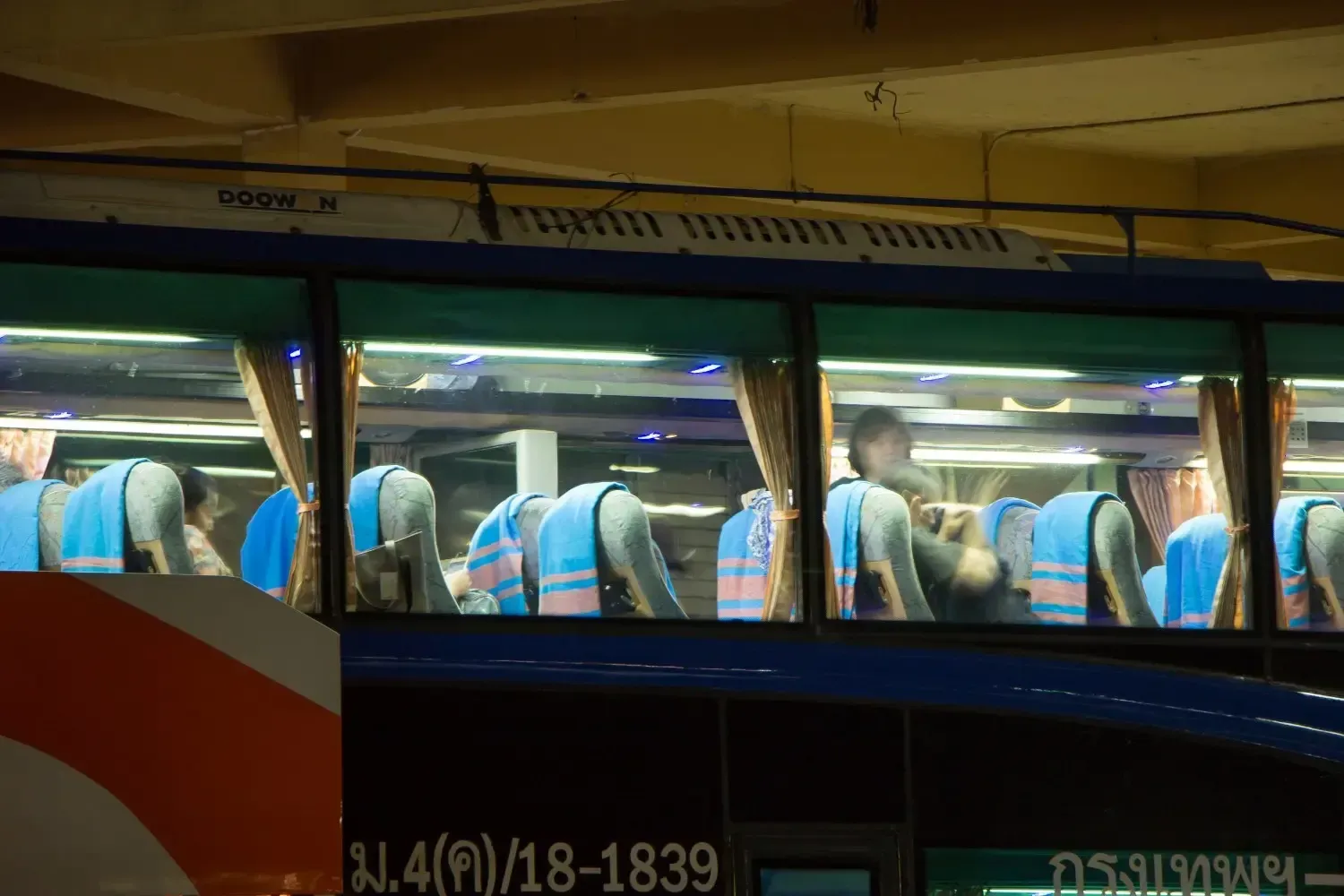 Interior de un autobús azul con pasajeros, iluminado de noche. Se ven las cortinas de las ventanas.