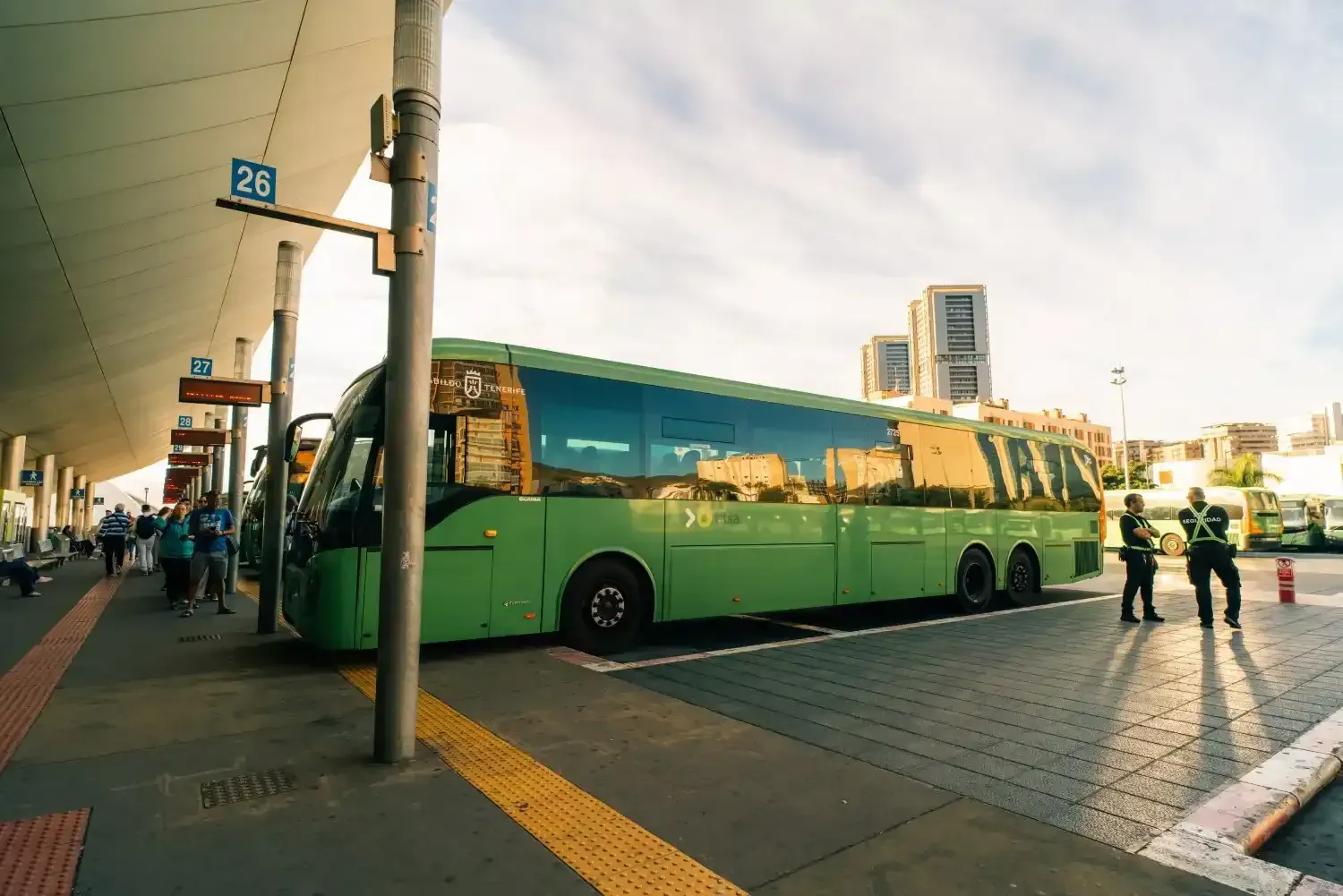 Autobús verde en una estación de autobuses. Gente esperando. Edificios al fondo.
