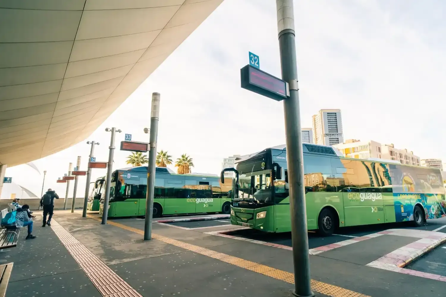 Autobuses verdes esperando en una estación de autobuses. La gente está sentada y caminando. El cielo está nublado.