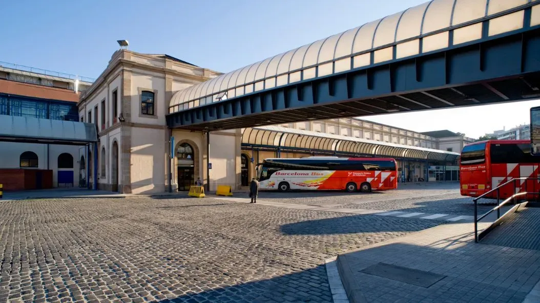 Puente peatonal cubierto sobre una estación de autobuses empedrada con autobuses rojos y un tren al fondo.
