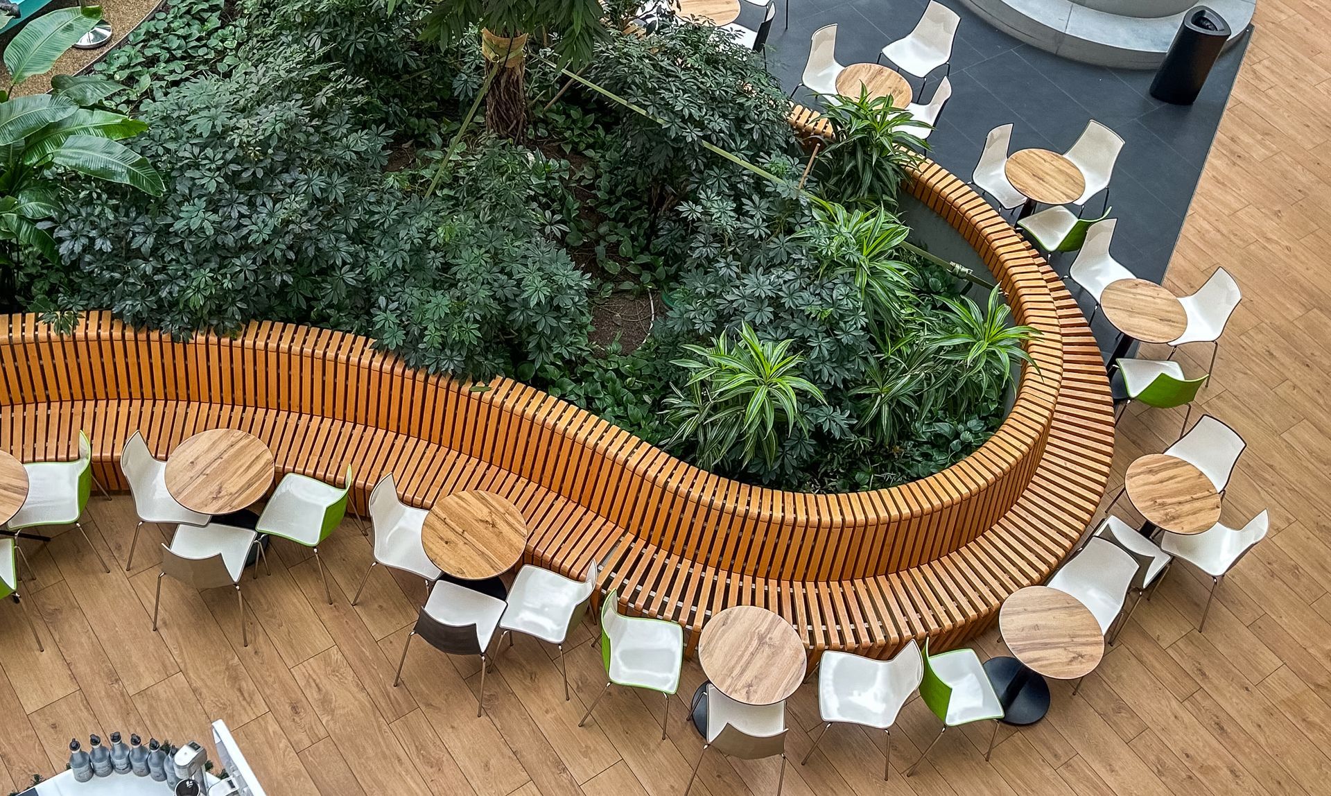 Jardinière en bois incurvée remplie de verdure dense, entourée de petites tables rondes et de chaises dans un atrium intérieur.