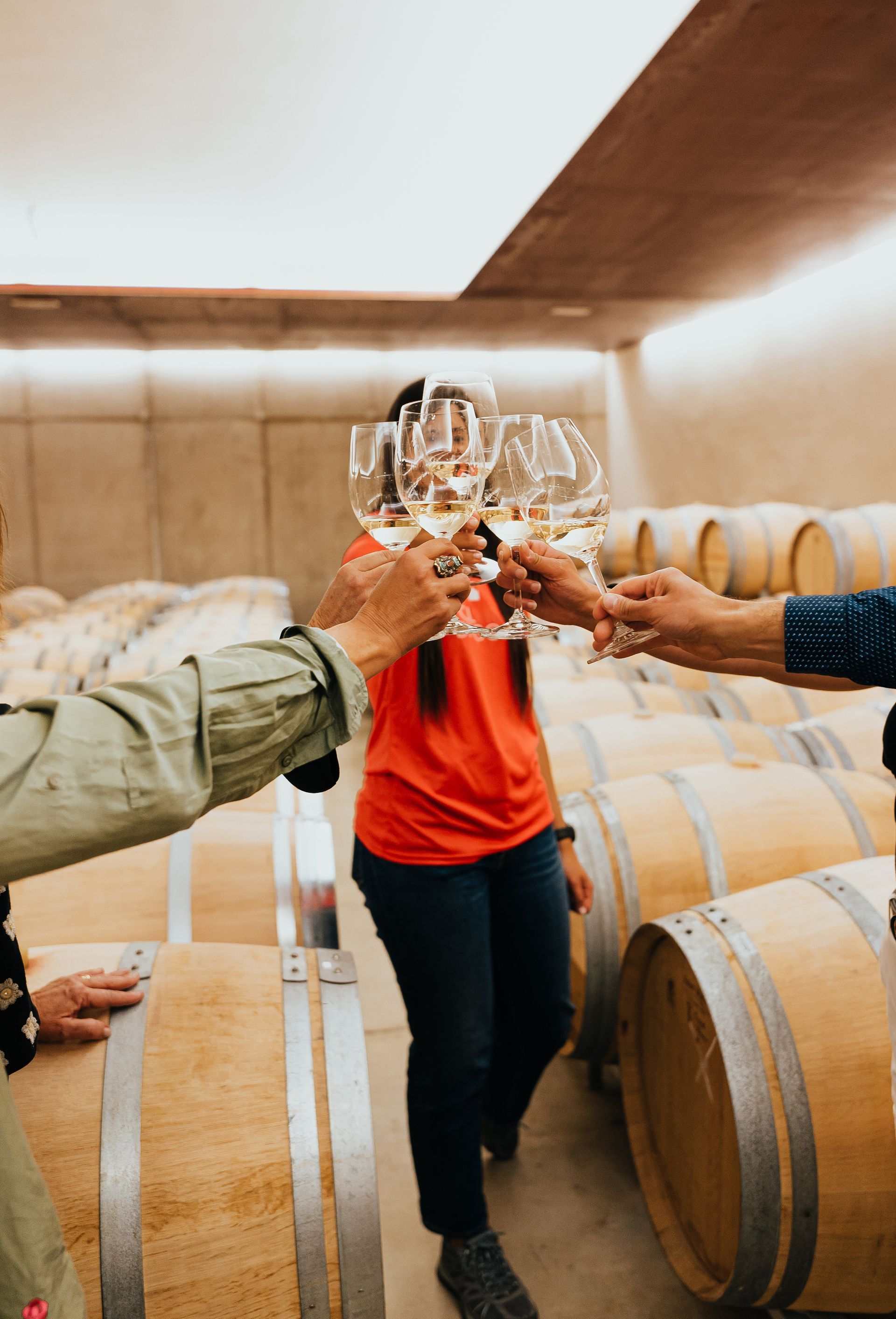 Des mains qui s'entrechoquent dans des verres à vin, avec des tonneaux en bois et une personne en chemise au centre.