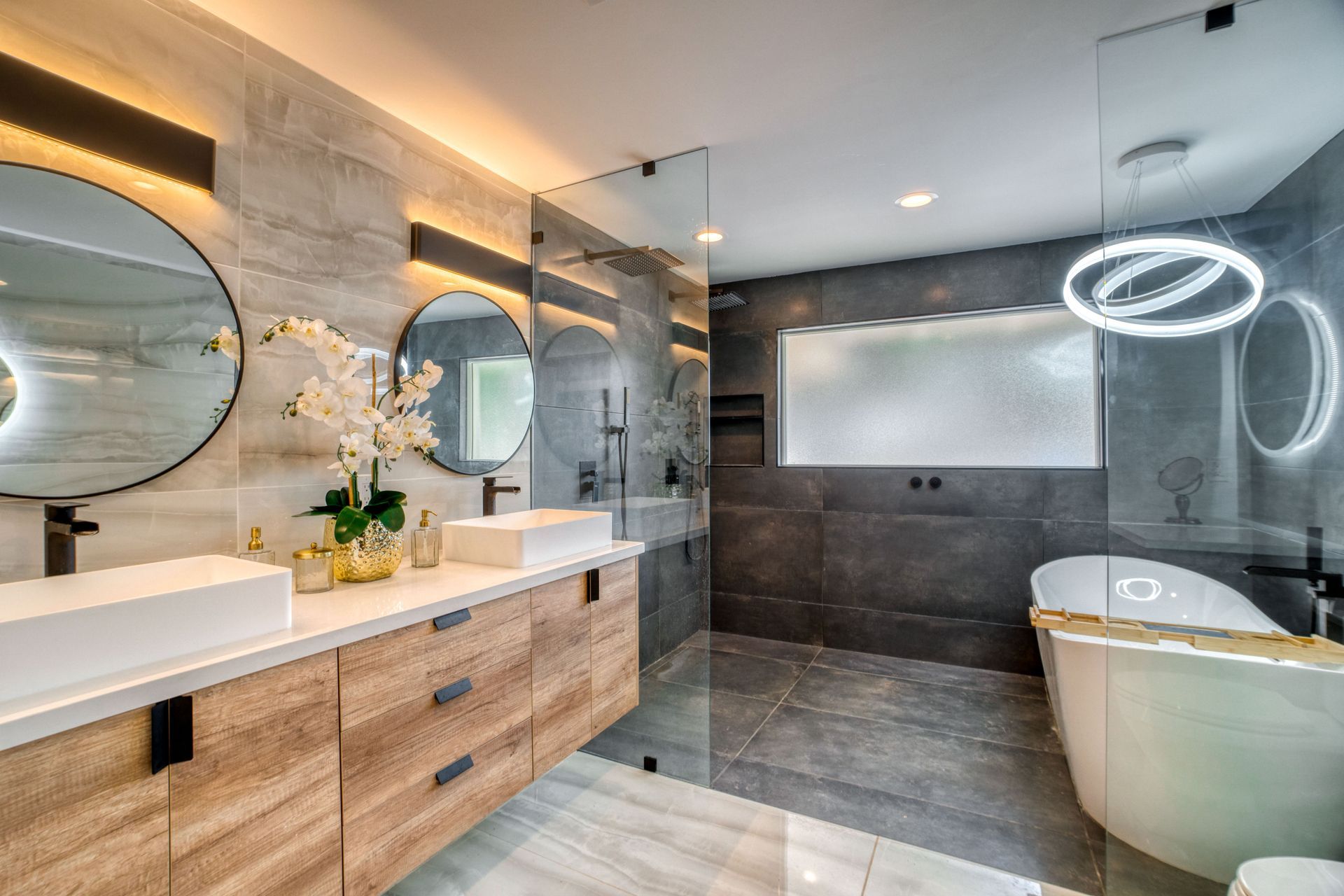 Salle de bains moderne avec double vasque, miroirs ronds, douche en verre et baignoire îlot sous un éclairage annulaire.