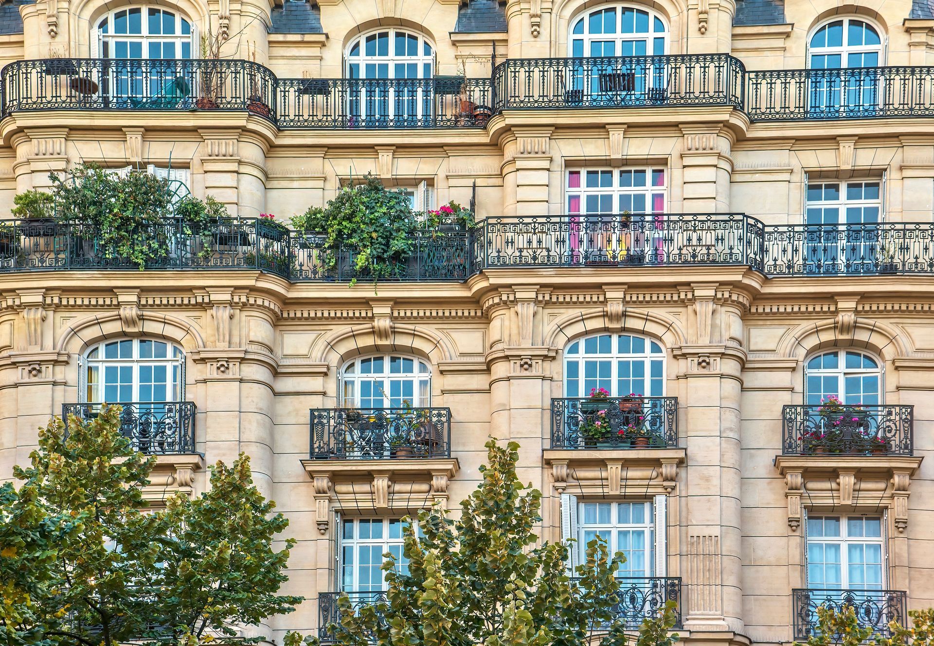 Immeuble parisien beige avec fenêtres cintrées, balcons en fer forgé et quelques espaces verts.