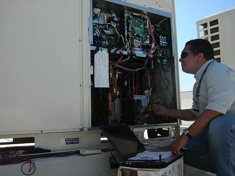 Un hombre está trabajando en un aire acondicionado con una computadora portátil.