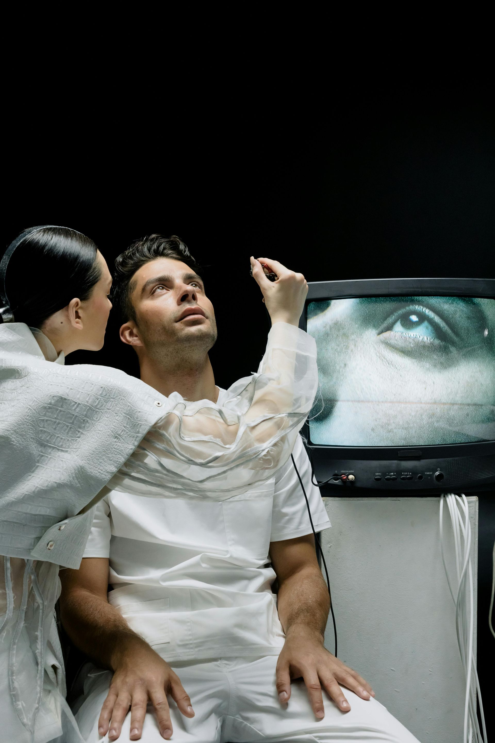 Uma mulher de jaqueta branca coloca algo no olho de um homem; uma TV com um olho está atrás deles.