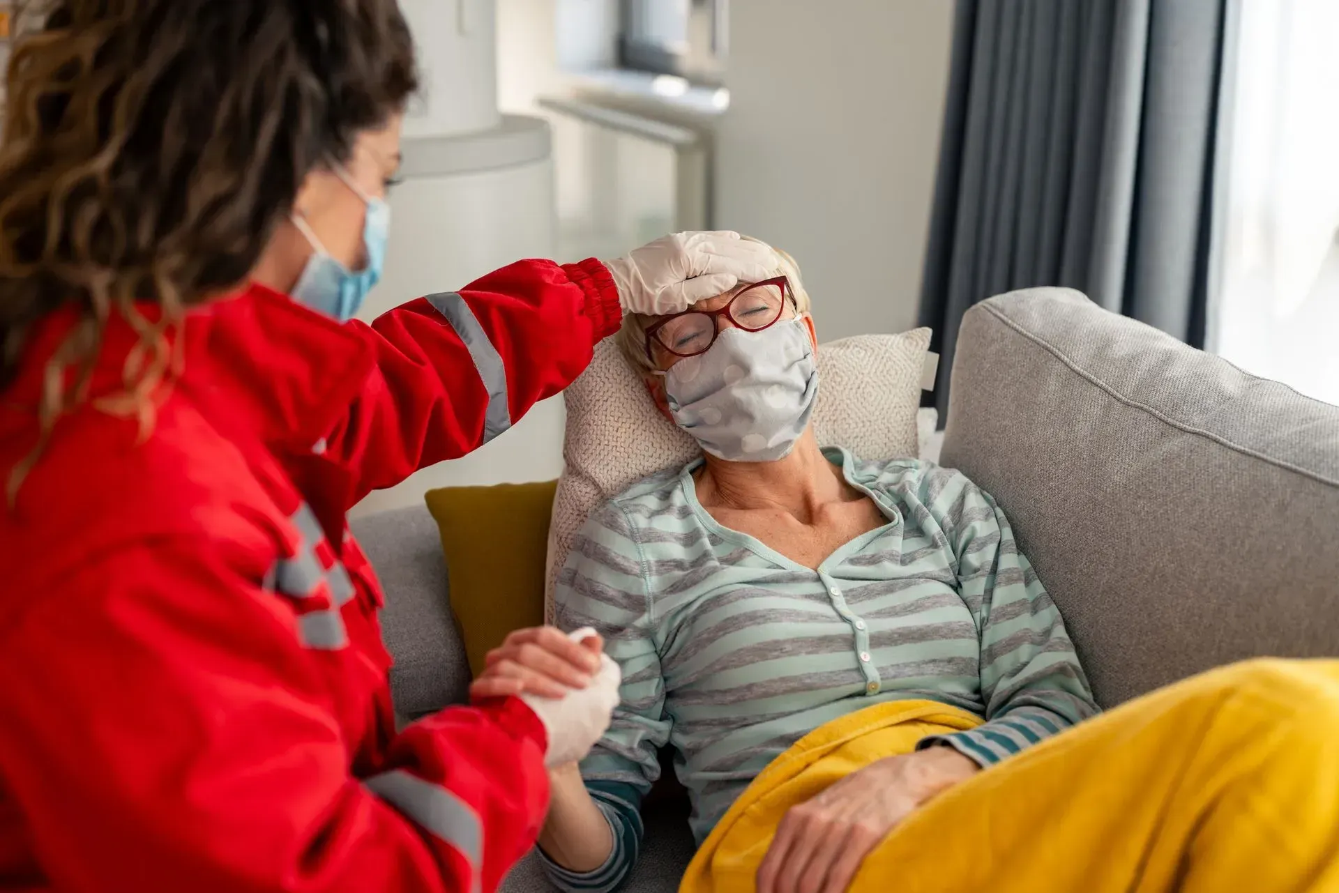 Un agent de santé vêtu d'une veste rouge examine le front d'une personne portant un masque et allongée sur un canapé.