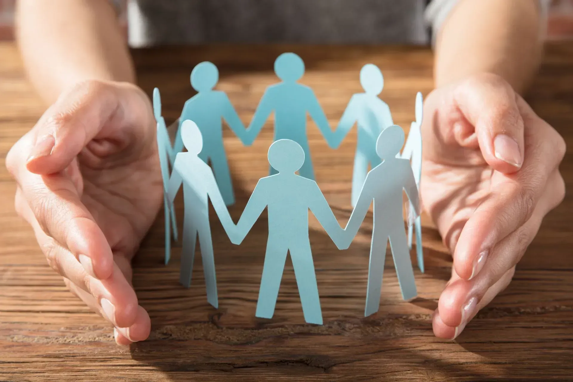 Deux mains enveloppent délicatement un cercle de personnes se tenant la main, découpé dans du papier, posé sur une table en bois.