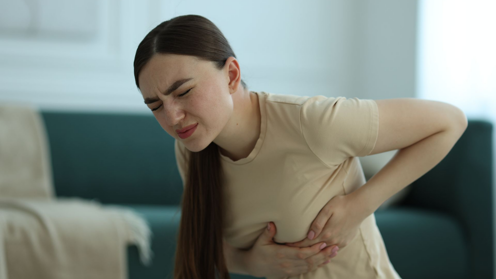 Une personne vêtue d'une chemise beige, assise dans un salon, se tient la poitrine avec une expression de douleur.