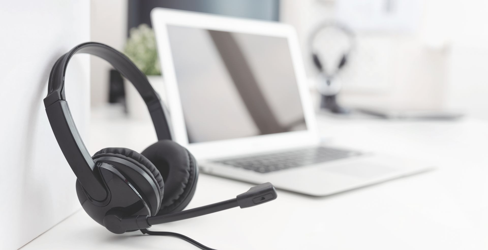 Un casque audio noir filaire avec microphone est posé sur un bureau blanc à côté d'un ordinateur portable dont l'image est floue.
