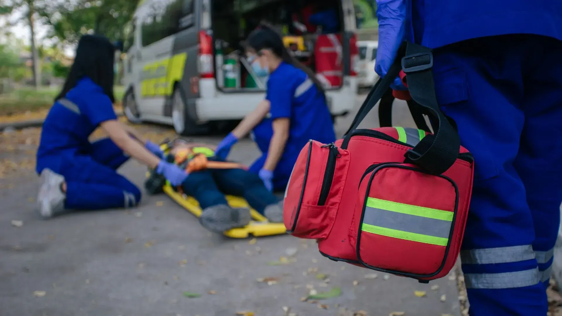 Des secouristes en uniforme bleu assistent une personne sur une civière près d'une ambulance, avec une trousse médicale au premier plan.