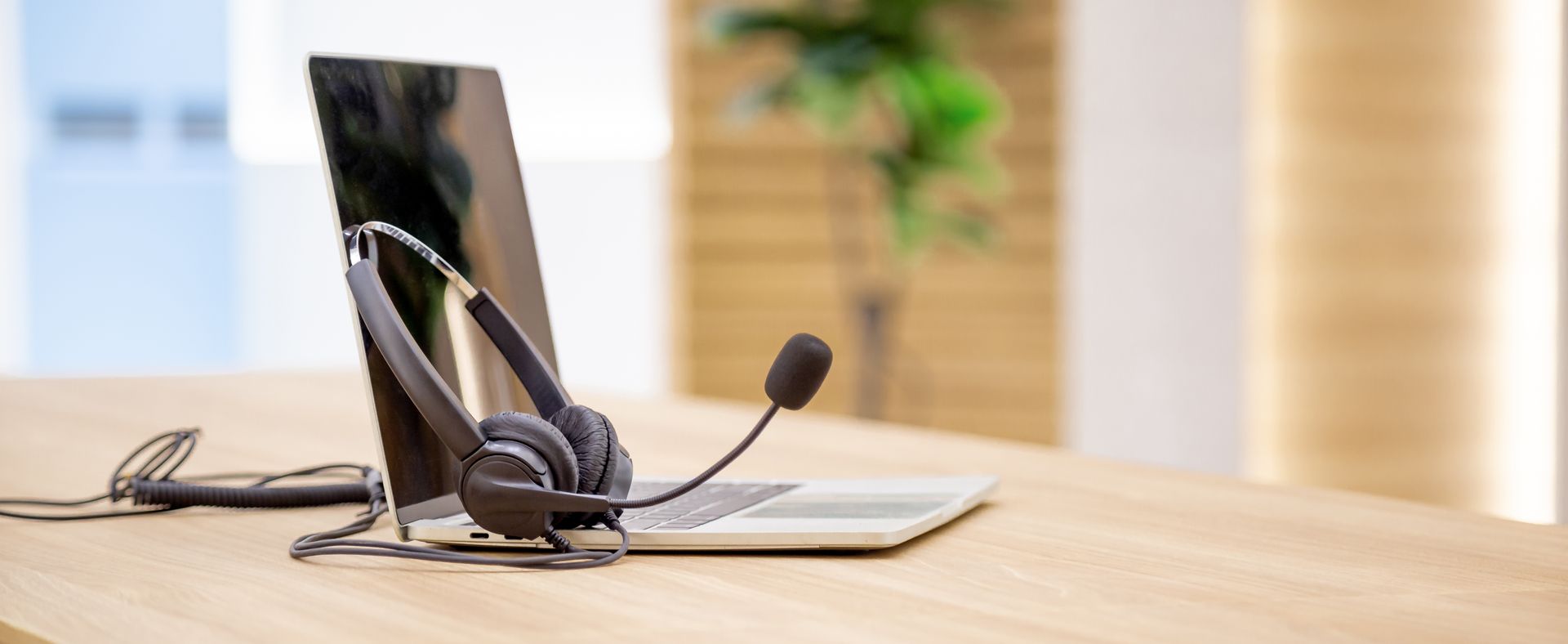 Dans un bureau lumineux, deux casques d'écoute noirs filaires avec microphone reposent sur un ordinateur portable argenté fermé, lui-même posé sur un bureau en bois clair.