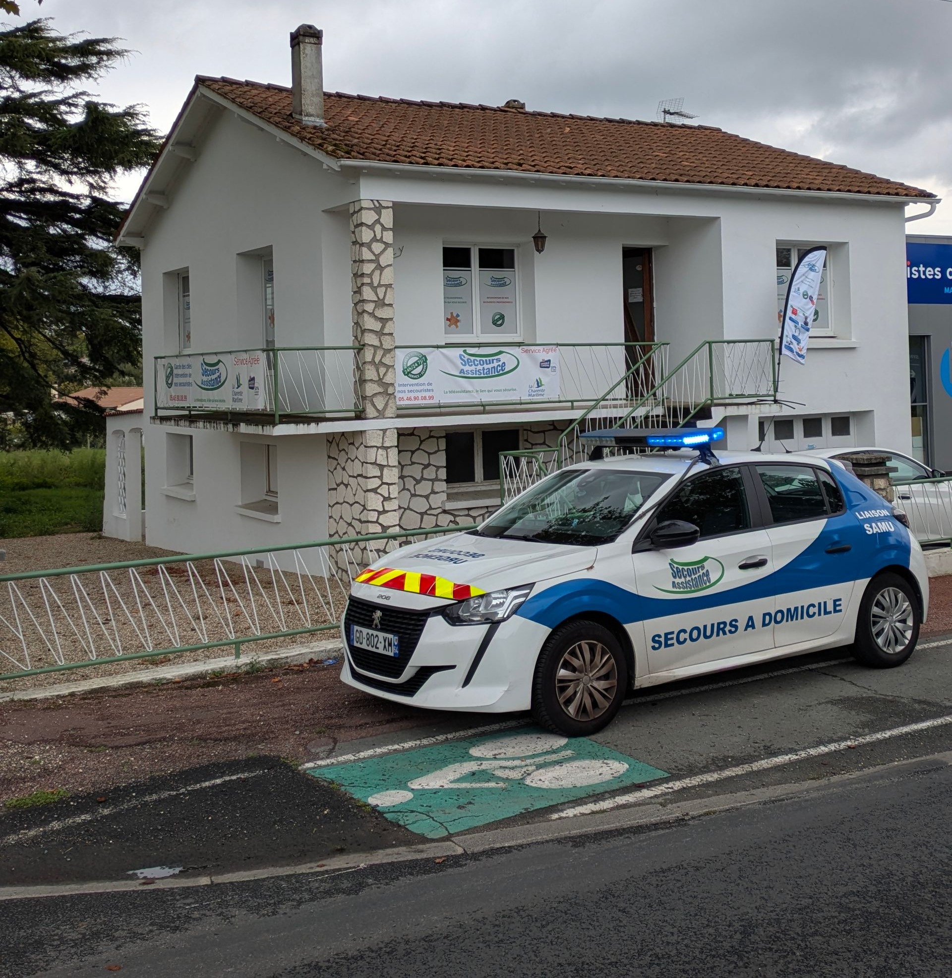 Une voiture de secours blanche et bleue était garée devant un bâtiment blanc de deux étages au toit de tuiles rouges.
