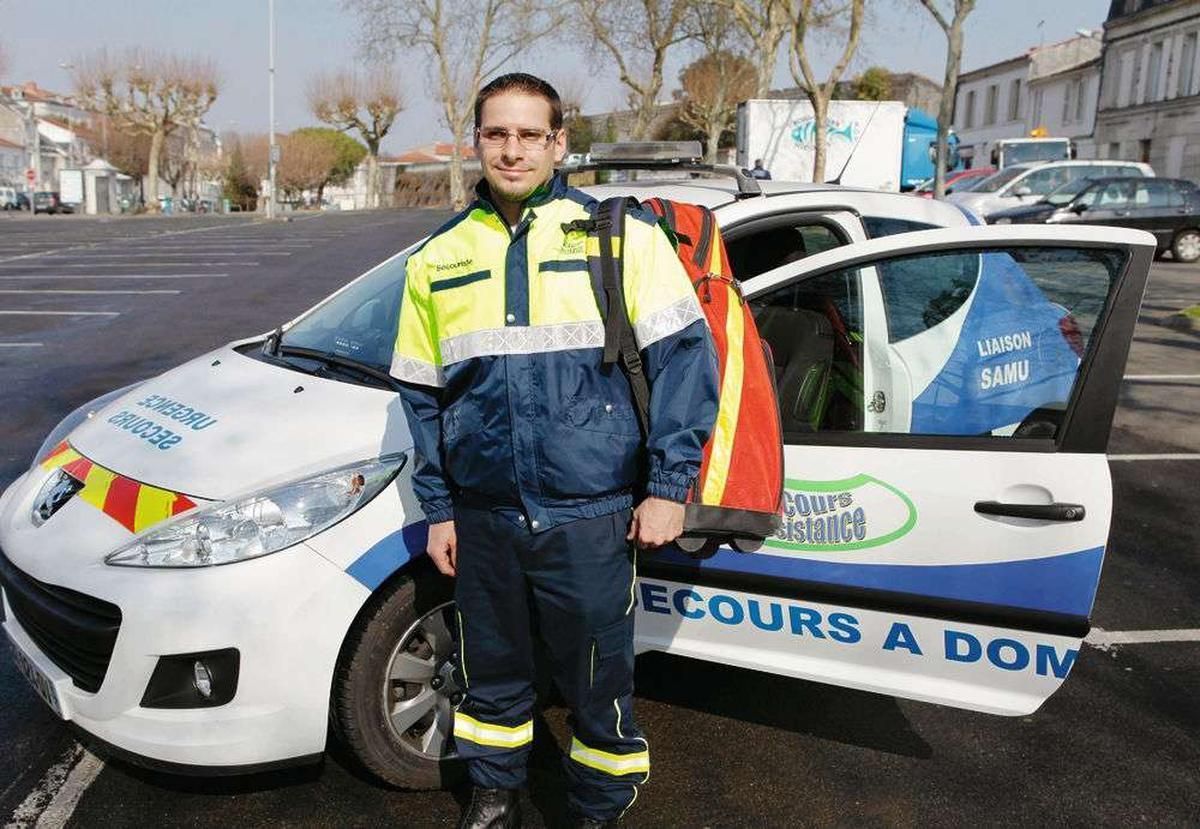 Un professionnel de santé en uniforme haut visibilité se tient à côté d'une voiture de service blanche portant l'inscription Sécurise un domicile.