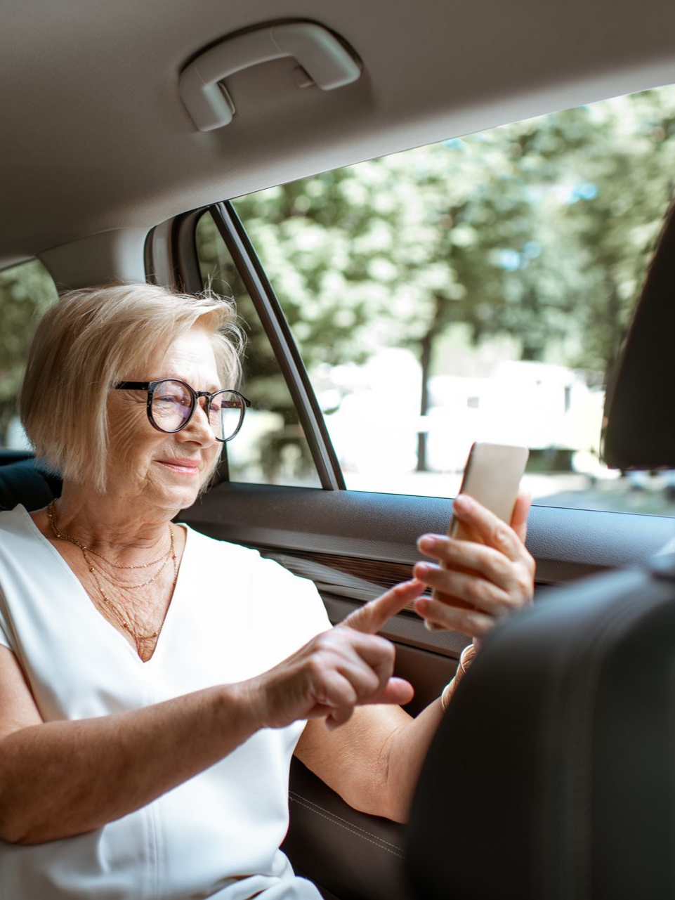 Femme dans une voiture portant des lunettes, regardant son téléphone, souriant, des arbres visibles à l'extérieur de la fenêtre.