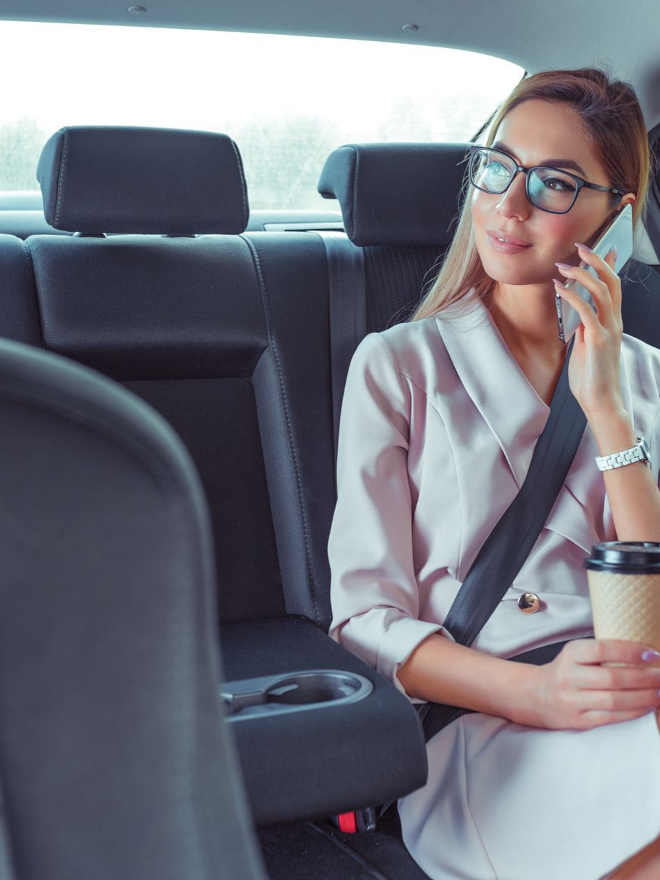 Femme en blazer rose avec un téléphone à l'oreille, assise sur la banquette arrière d'une voiture et tenant une tasse de café.