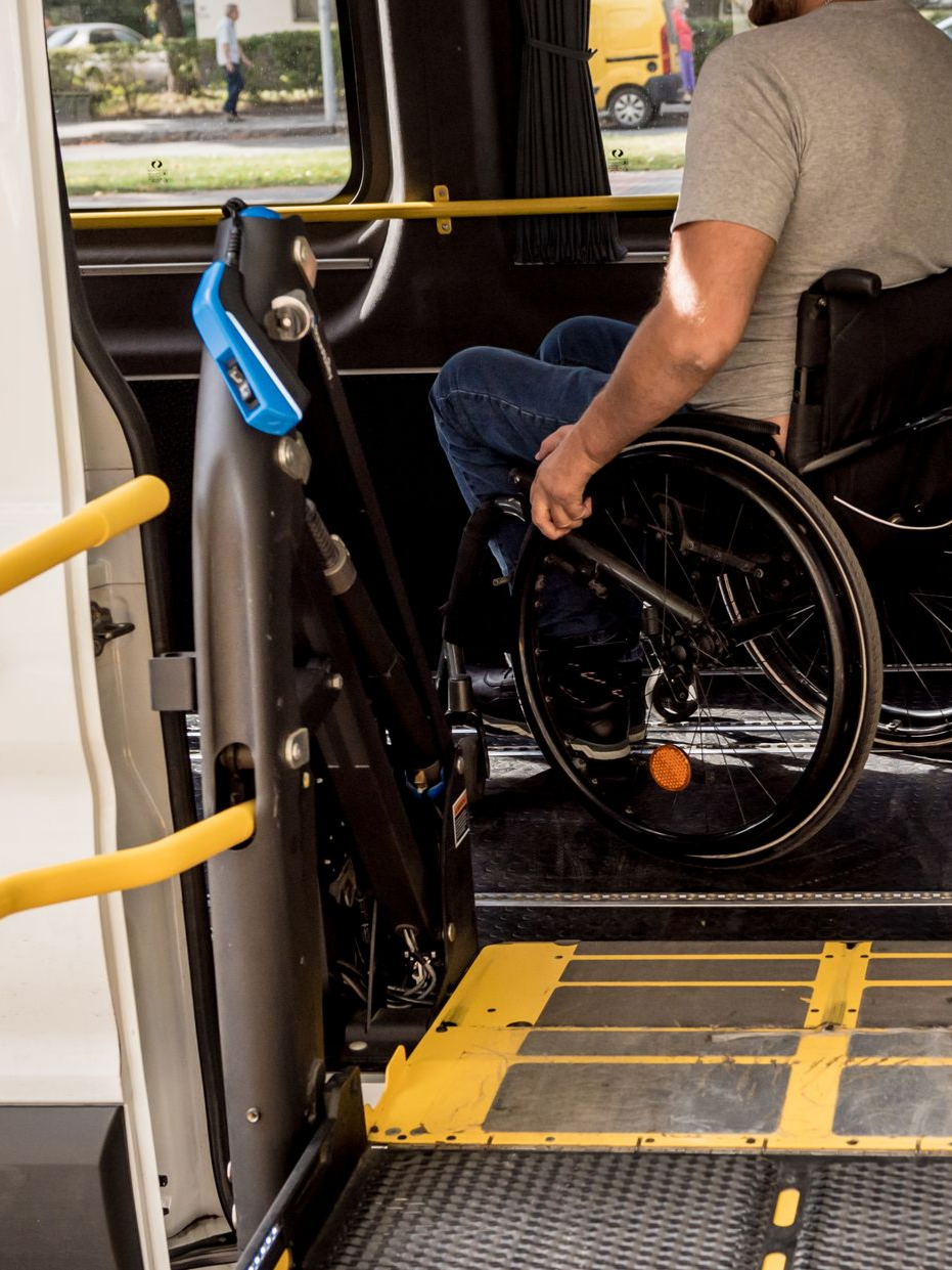 Homme en fauteuil roulant sur une rampe d'accès pour monter dans un bus. Barres de sécurité jaunes, commandes bleues.