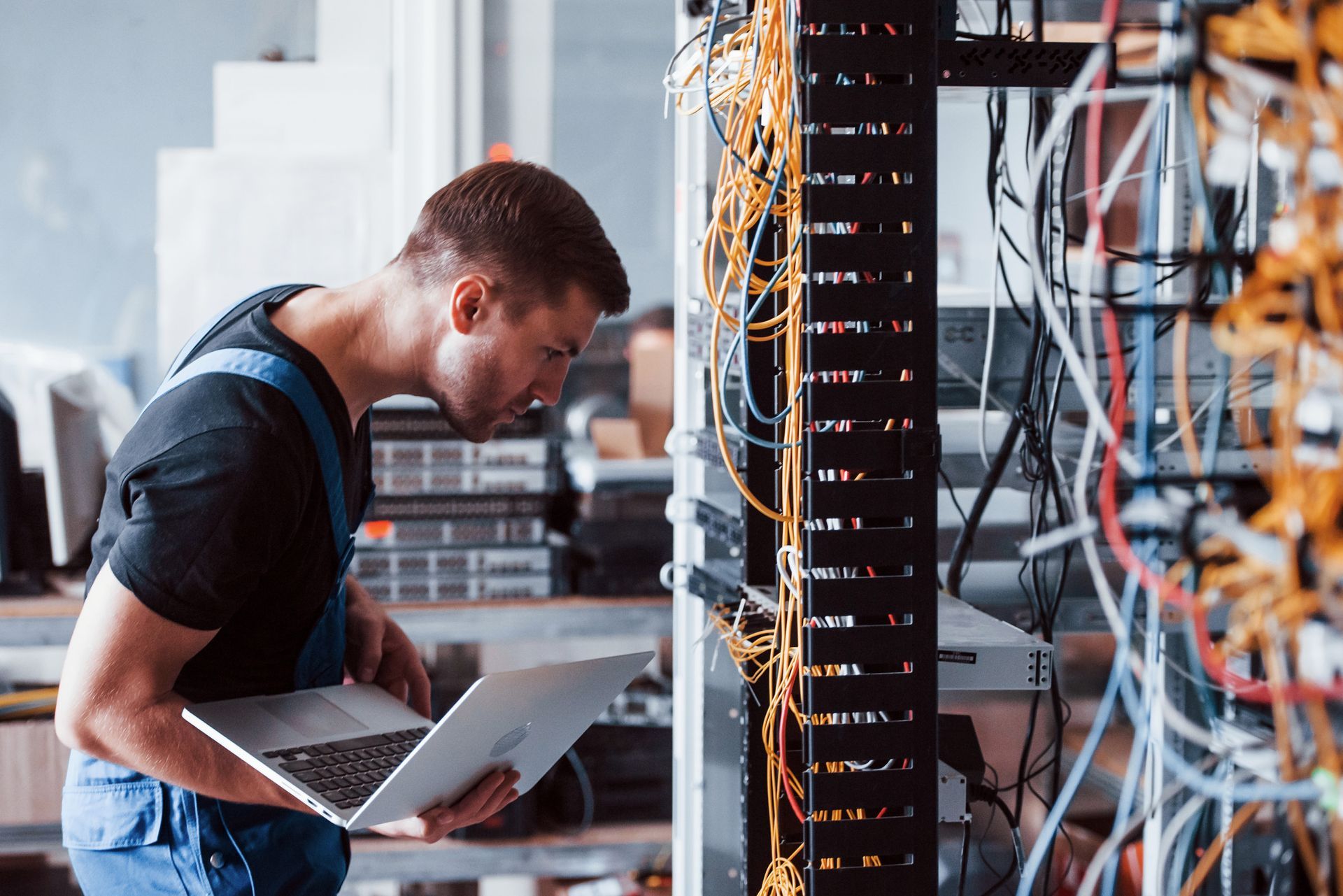 Hombre usando una computadora portátil, trabajando en un rack de servidores lleno de cables en un centro de datos.