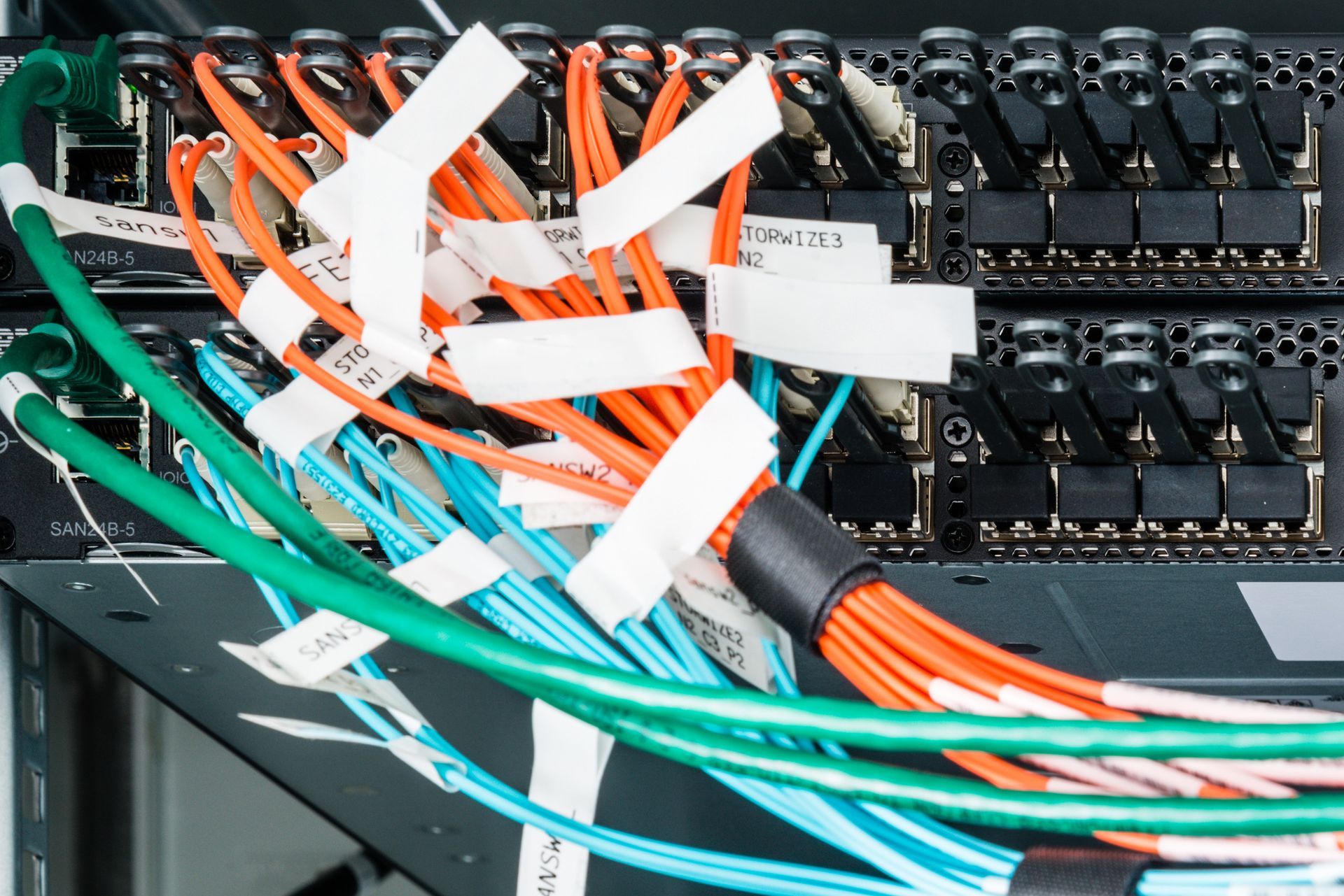Primer plano de cables de red conectados a un servidor, etiquetados con cinta; incluye cables naranja, verde y azul.