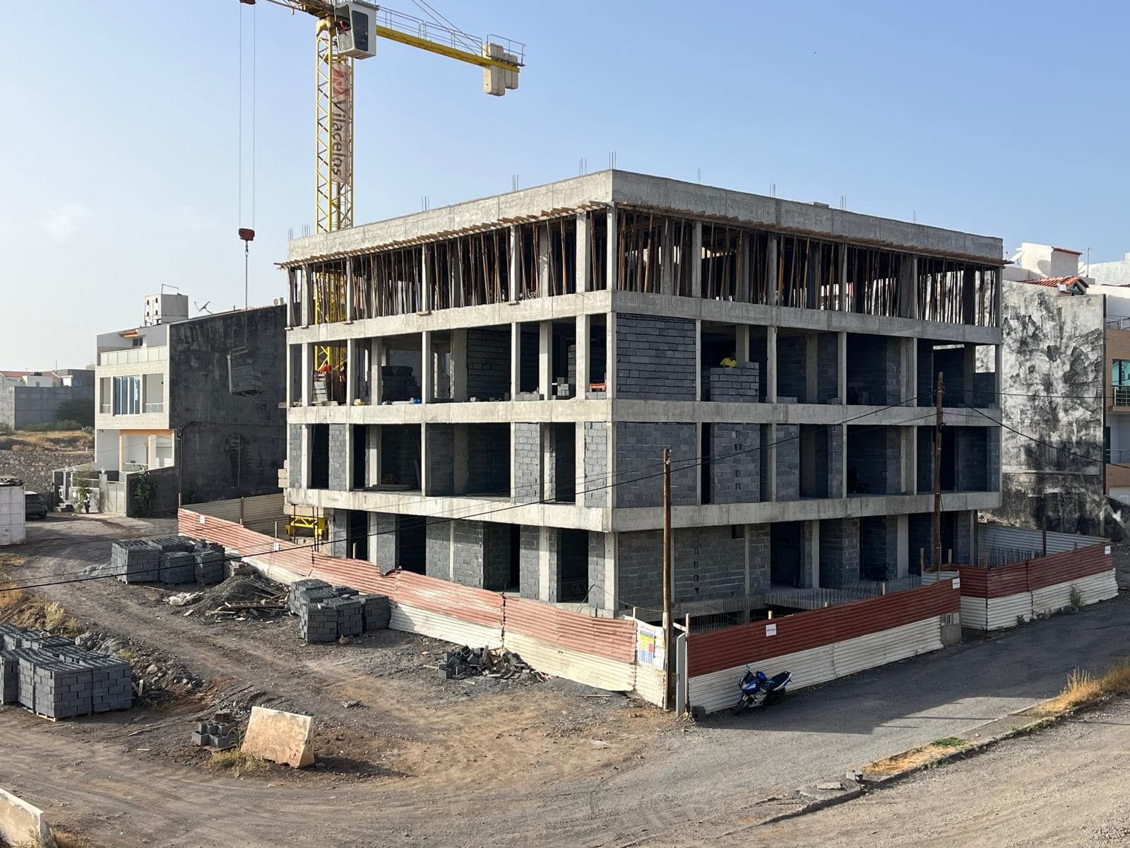 Prédio em construção com guindaste, cercado por entulho e um muro de tijolos.