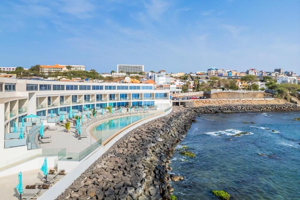 Resort costeiro com piscina turquesa, vista para o oceano e edifícios brancos de vários andares sob um céu azul.