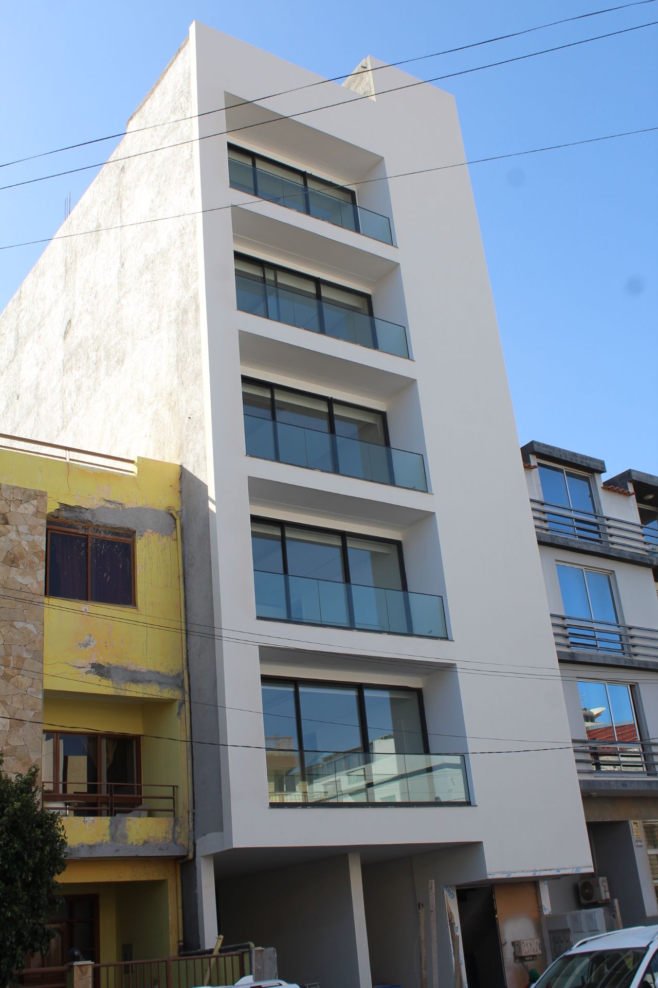 Prédio de apartamentos moderno e branco com varandas de vidro, em uma rua com outros prédios.